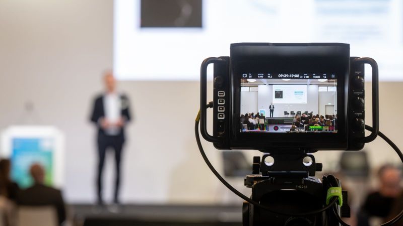 Vortragender auf der Bühne im Hintergrund, im Vordergrund die Kamera, die ihn dabei filmt.
