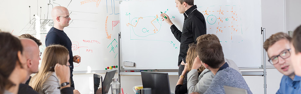 Personen mit PC und Notizblöcken am Tisch blicken auf beschriftetes Board, an dem einer der Gruppe etwas erklärt