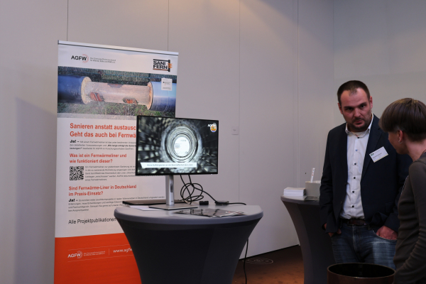 Zwei Teilnehmende stehen am Stand des Projekts SaniFern. Ein Bildschirm zeigt eine Innenansicht einer sanierten Fernwärmeleitung; dahinter informiert ein Aufsteller über das Verfahren zur grabenlosen Sanierung.
