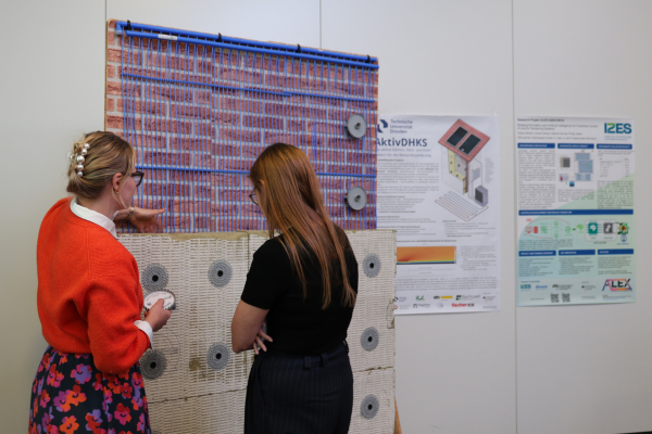 Zwei Teilnehmerinnen betrachten eine Beispielwand des Projekts AktivDHKS. Auf der Wand sind Rohrleitungen und Befestigungselemente der entwickelten Sanierungslösung sichtbar.