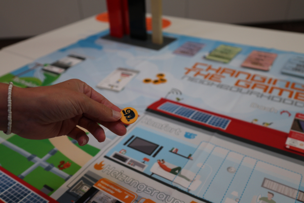 Eine Hand hält einen gelben „Fauli“-Spielchip über dem Brettspiel „Changing the Game – Neighbourhood“. Auf dem Spielbrett sind Gebäude, Energieanlagen und weitere Elemente eines Stadtquartiers dargestellt.