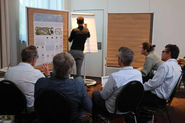 Mehrere Teilnehmer sitzen in einem Workshopraum vor einem Flipchart. Ein Teilnehmer steht am Flipchart und notiert Ergebnisse der Gruppenarbeit.