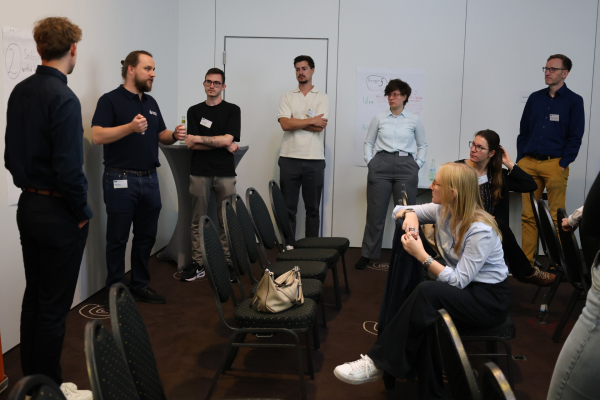 Teilnehmende eines Workshops stehen und sitzen in einem Veranstaltungsraum. Ein Teilnehmer spricht zur Gruppe; an den Wänden hängen beschriftete Arbeitsblätter aus der Gruppenarbeit.